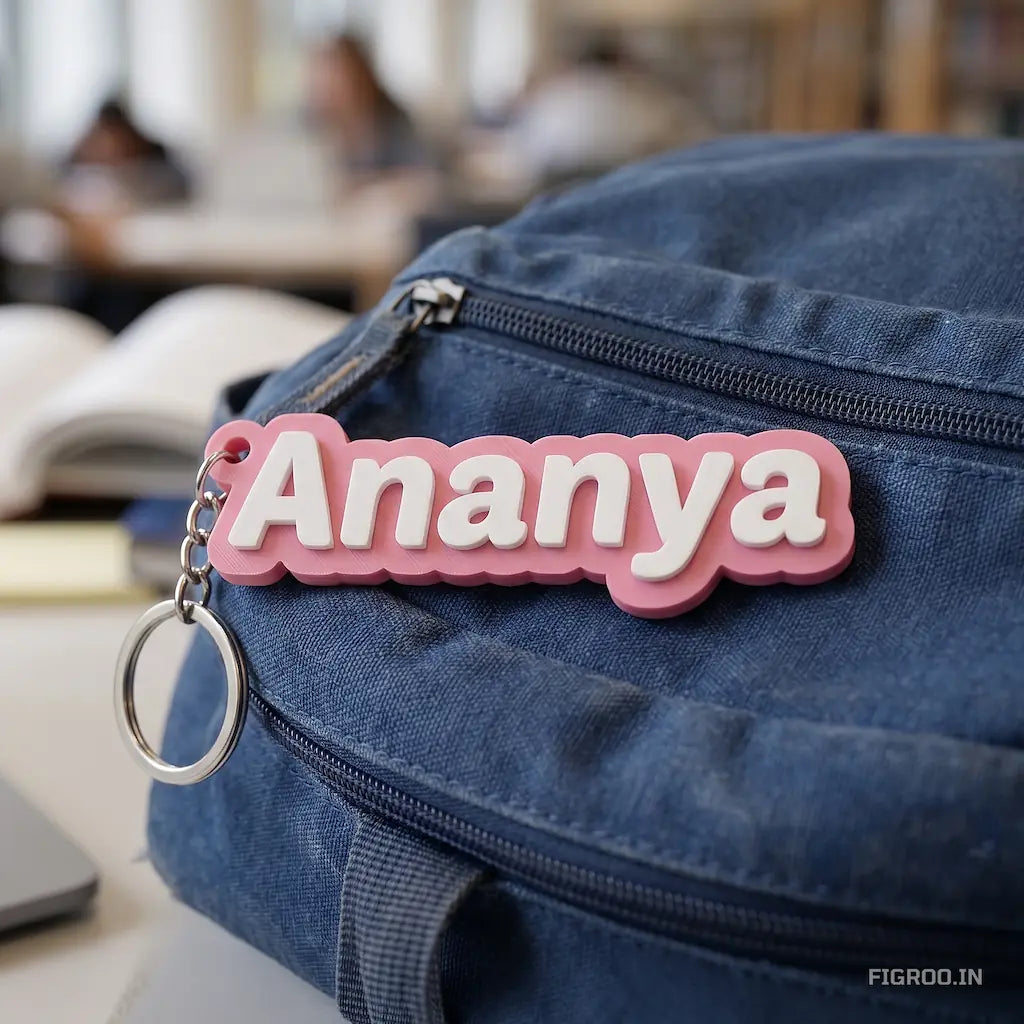 A personalized 3D printed name tag spelling Ananya with a pink base and white text, attached as a zipper pull to a blue canvas backpack.